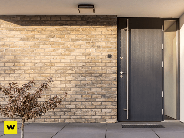 Moderne Haustür aus dunklem Material in Klinkerfassade mit Vordach und Pflanzen.