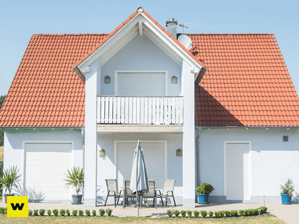 Modernes Einfamilienhaus mit rotem Dach, Balkon und Terrasse, Gartenmöbel im Vordergrund.